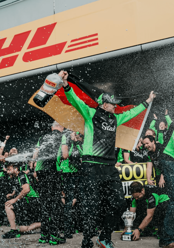 Nico Hülkenberg holds a German flag behind his head as he is sprayed with Champagne by his Sauber team. In his right hand, he is holding a bottle of Champagne and a Pirelli cap.