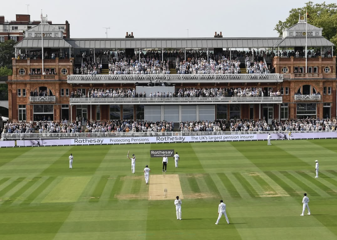 This photo shows a view of the prestigious old grandstand at Lord's Cricket Ground. The view shows one batsman raising his bat, whilst fielders stand in their positions.