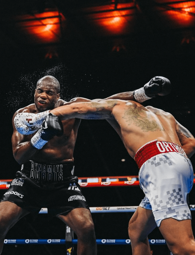 This photo shows Usyk landing an overhand left punch on Dubois. There is a spray of sweat around Dubois's head and neck. Dubois is wearing black trunks and Usyk is wearing white and red.