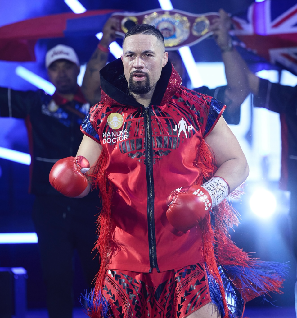 This image shows Joseph Parker during a recent ringwalk. He looks focused and is wearing a red robe and red gloves. This team carry his belt in the background.