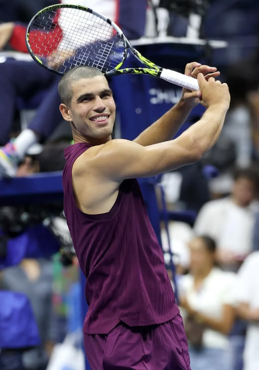 This photo shows Carlos Alcaraz grinning whilst holding his tennis racket behind his head.